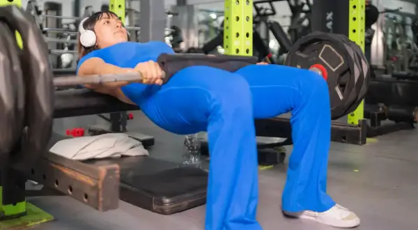 Kobieta w niebieskim stroju wykonuje hip thrust, jedno z najlepszych ćwiczeń na pośladki, z obciążeniem na sztandze.